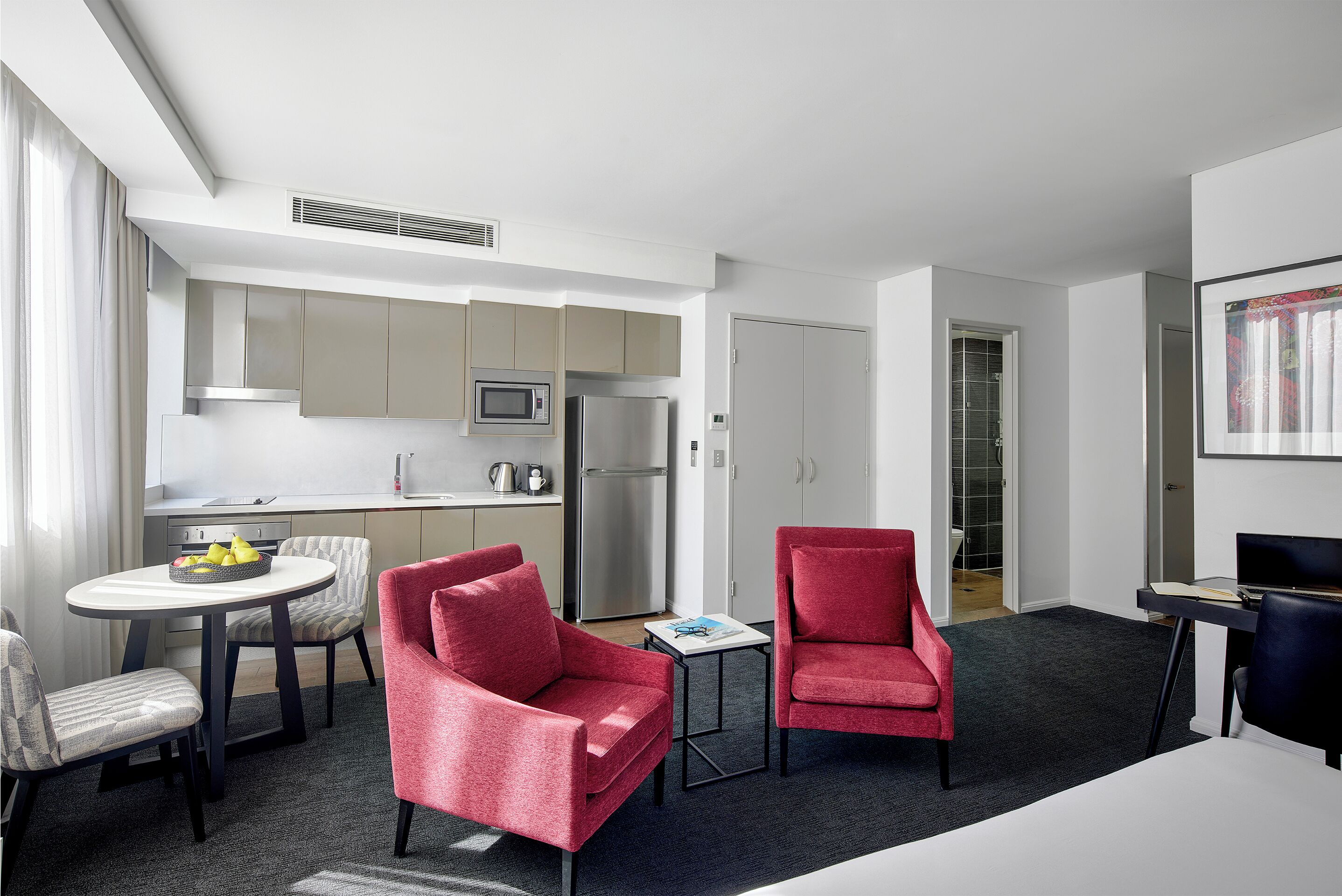 A modern hotel room with a kitchen, two red chairs, a small table, and a desk with a computer.