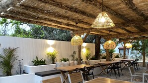 Un patio al aire libre con mesas y sillas de madera, lámparas de mimbre colgantes y plantas en macetas.