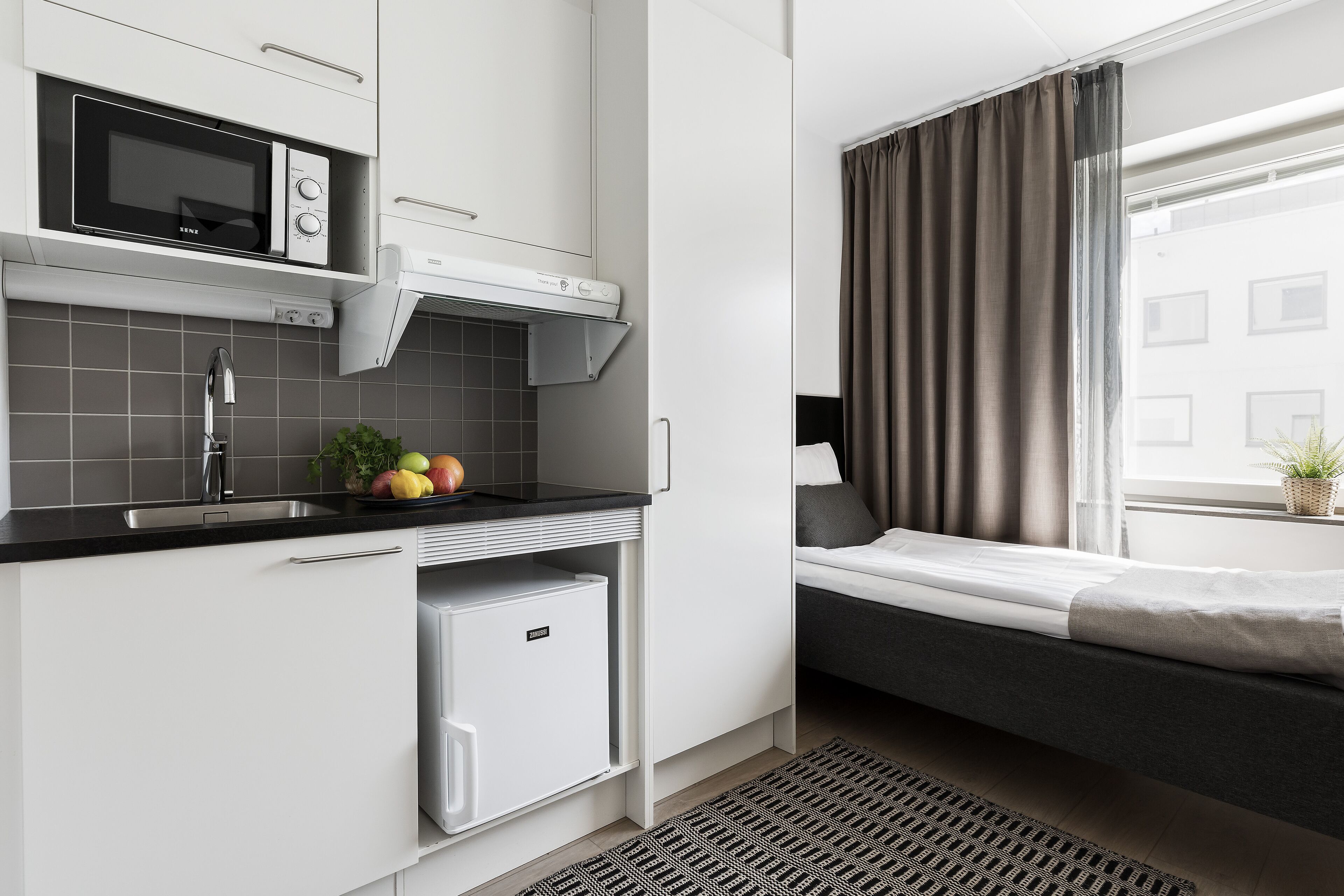 A compact kitchen with a microwave, refrigerator, and sink. A small dining area with a bench and a window with curtains.