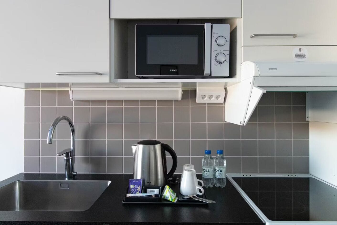 A modern kitchen with a stainless steel sink, a kettle, a microwave, and a coffee maker. There are water bottles and a packet of tea on the counter.