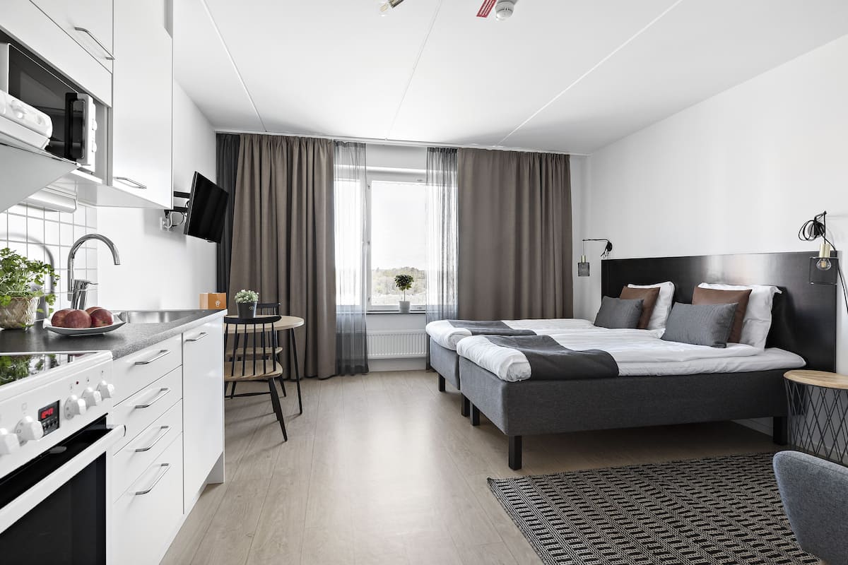 Une chambre d’hôtel moderne dotée d’une kitchenette, d’un lit avec une literie grise, d’une petite table à manger et offrant une vue sur l’extérieur.