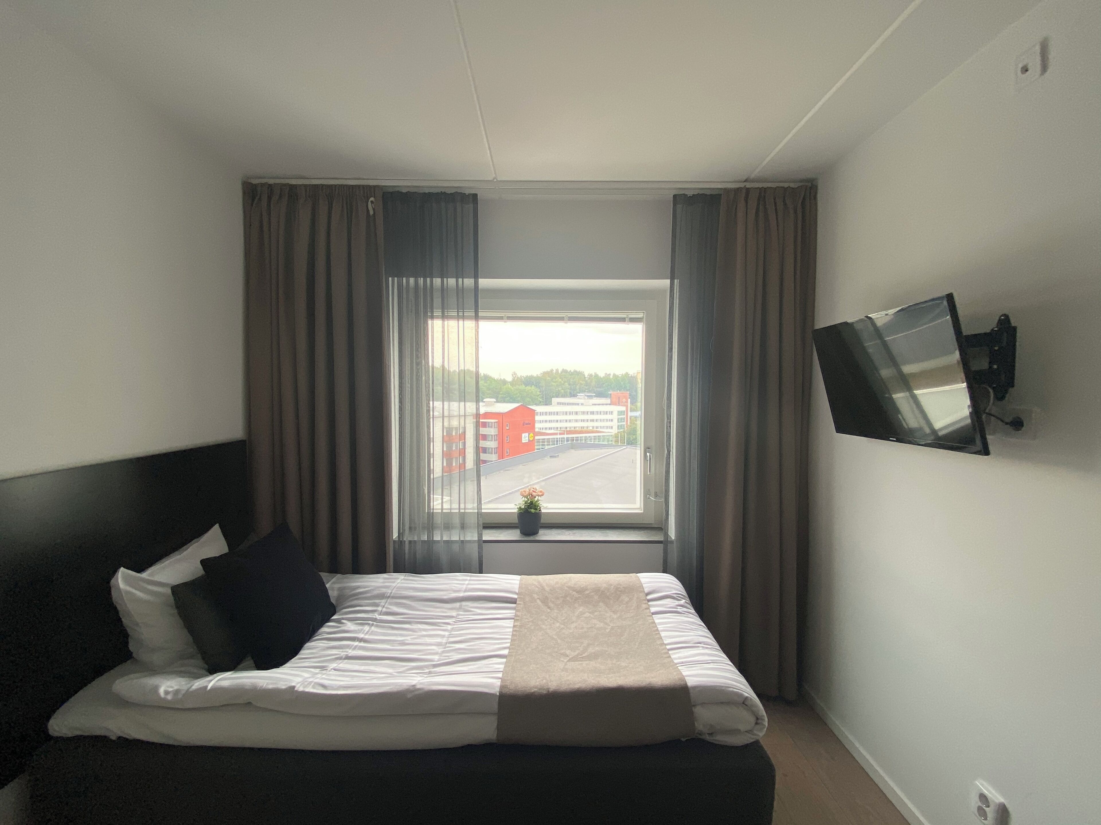 Een hotelkamer met een bed, gordijnen, een aan de muur bevestigde televisie en een raam met uitzicht op een parkeerplaats en gebouwen.