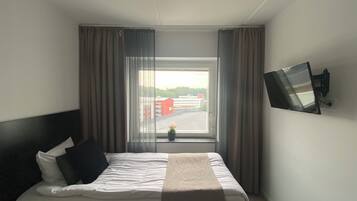 Een hotelkamer met een bed, gordijnen, een aan de muur bevestigde televisie en een raam met uitzicht op een parkeerplaats en gebouwen.