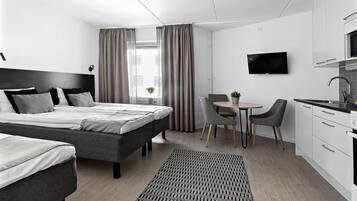 A modern hotel room with a large bed, a desk, a chair, a TV, and a kitchenette.
