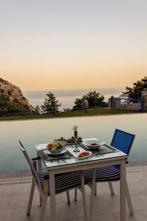 Une table à manger avec des assiettes remplies de nourriture, un vase et un ordinateur portable, le tout installé sur une terrasse offrant une vue sur la mer et un paysage rocheux.