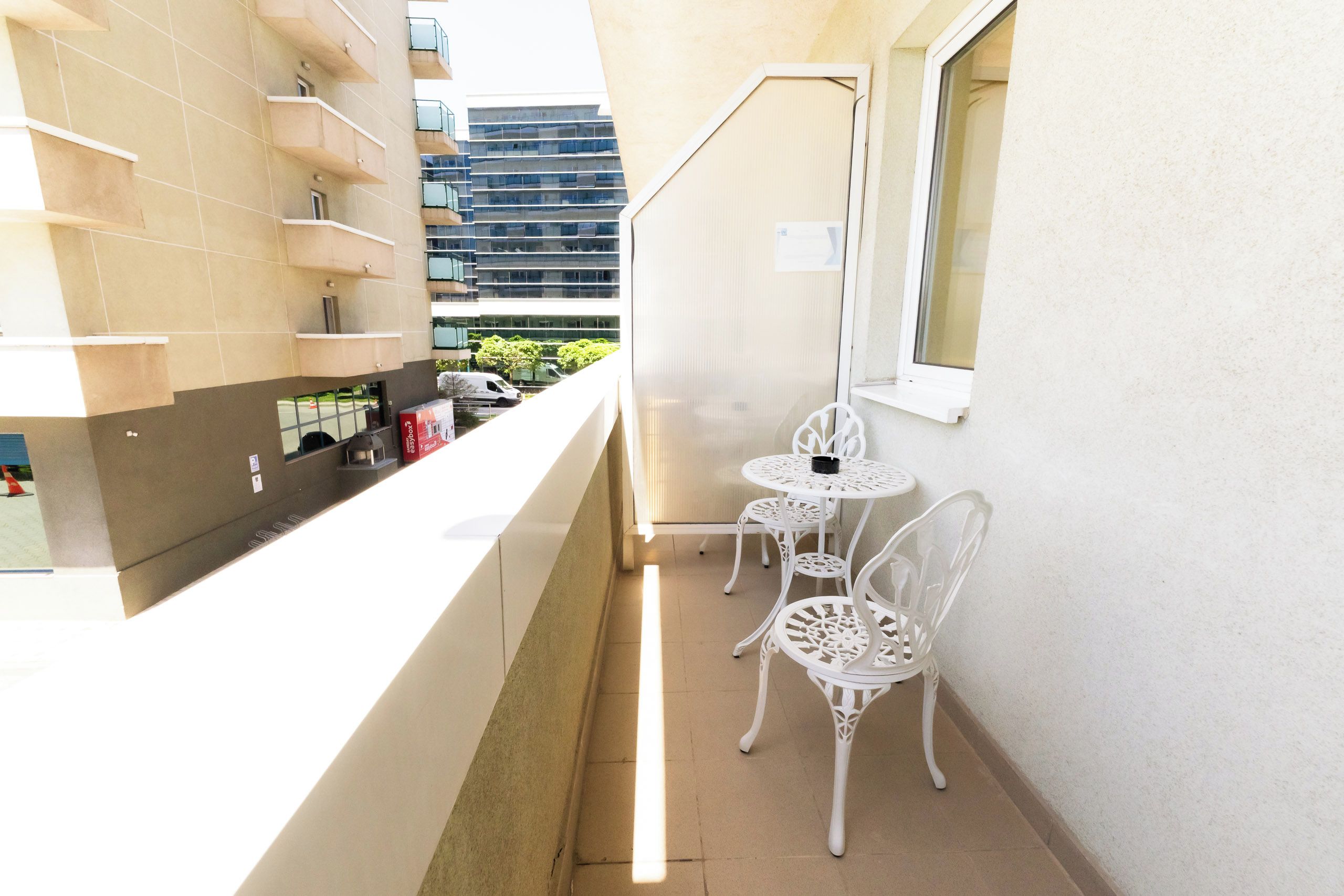 A narrow balcony with a kitchen area, two white metal chairs, and a window.