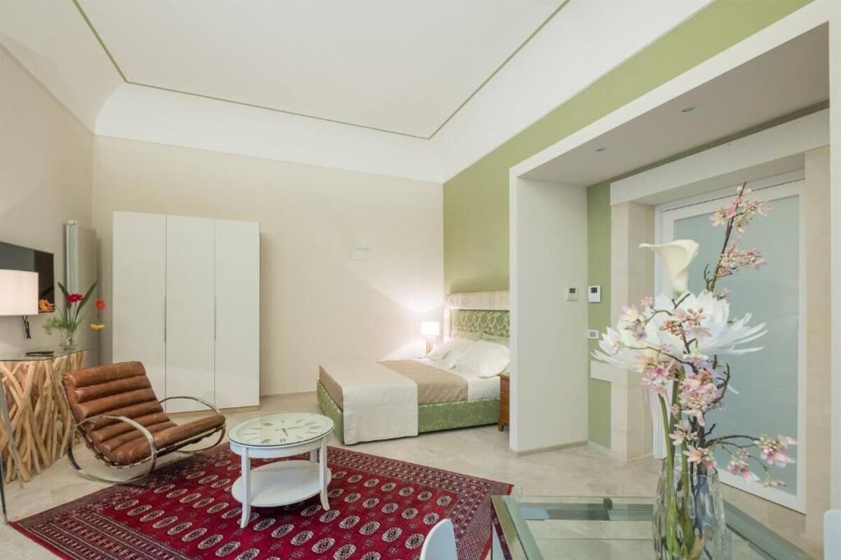 A hotel room with a bed, a chair, a small table, a red patterned rug, and a vase with flowers.