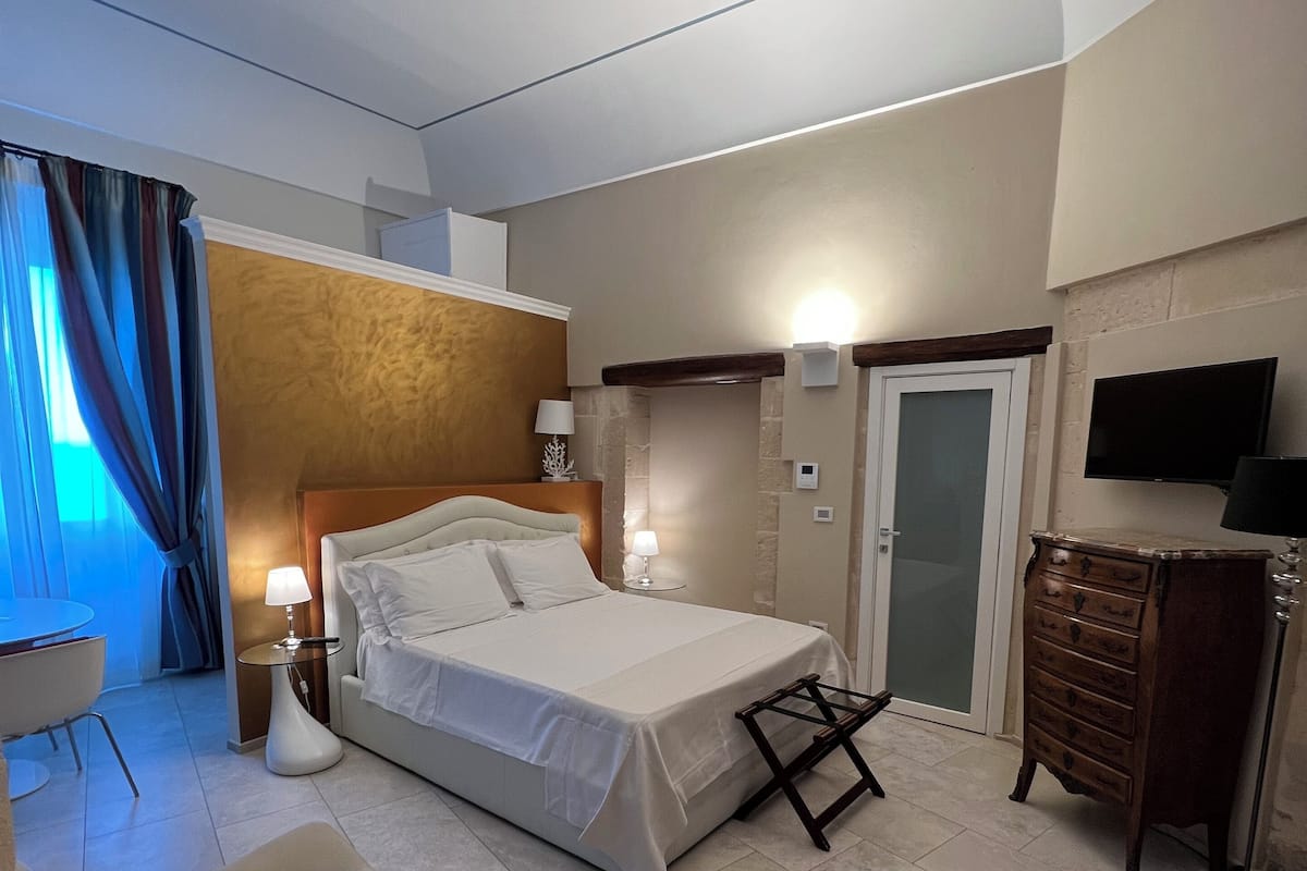 A modern hotel room with a large bed, a sitting area with a chair, a dresser, and a flat-screen TV mounted on the wall.