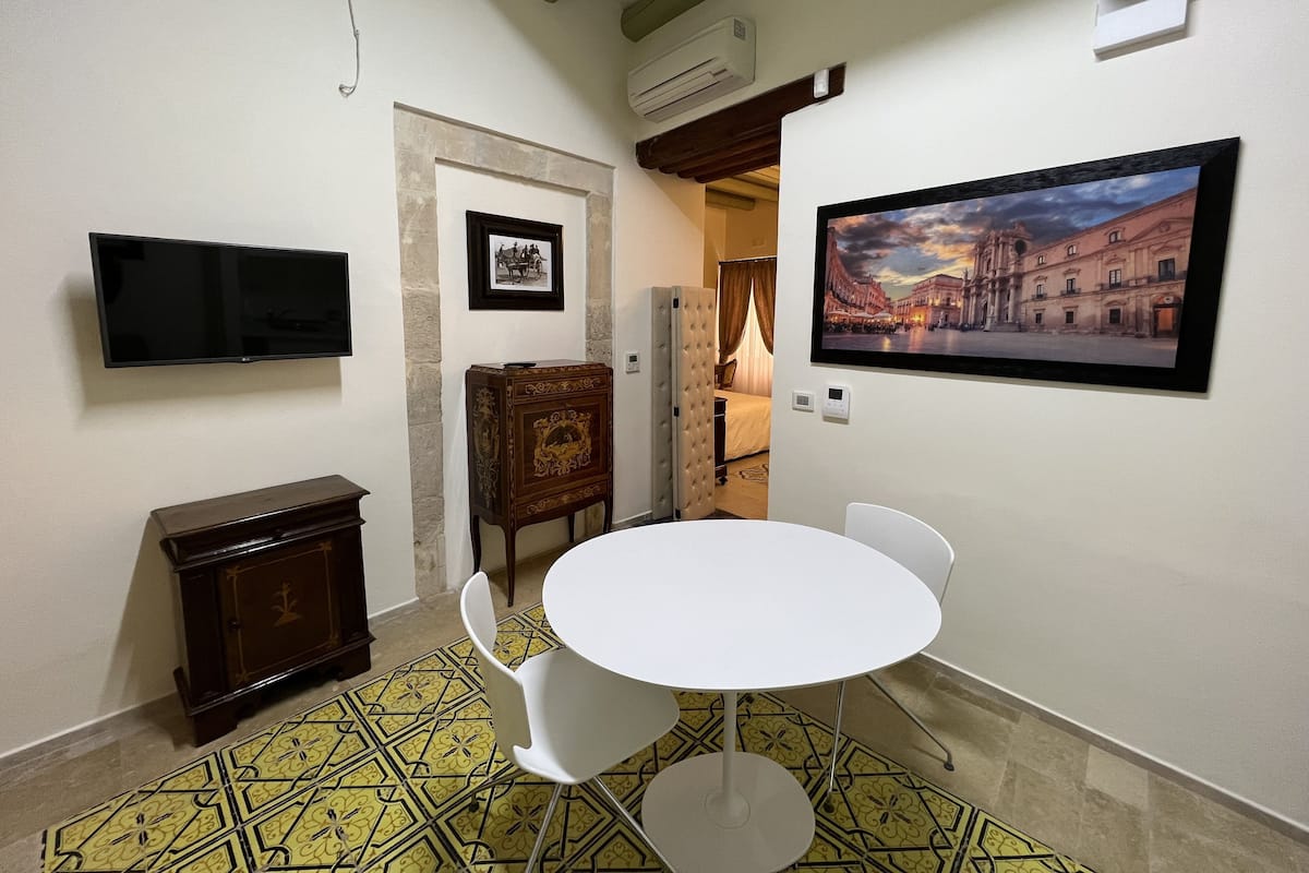 A hotel room with a round table, white chairs, a flat-screen TV, a framed picture, a wooden cabinet, and a patterned floor.