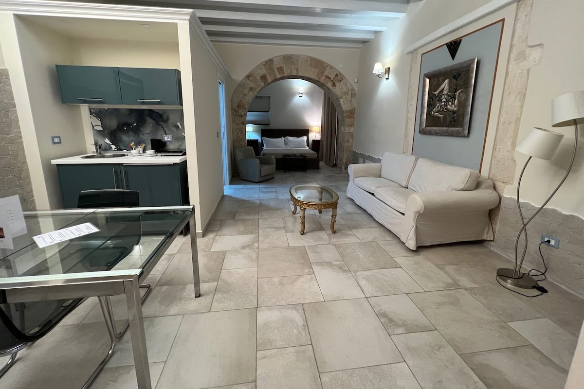 A modern living room with a glass table, a sofa, and a coffee table. There is a kitchen area with cabinets and a countertop. An archway leads to a bedroom with a bed and a nightstand.