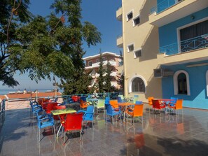 Un área de café al aire libre con sillas y mesas de colores, vistas despejadas al mar y un edificio con balcones al fondo.