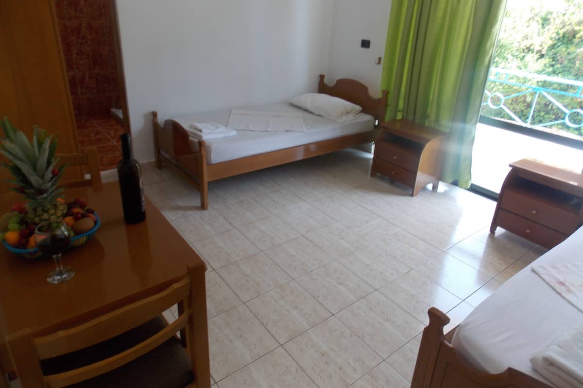 A bedroom with a bed, a wooden nightstand, a table with a fruit bowl and a bottle, and a view of greenery outside.