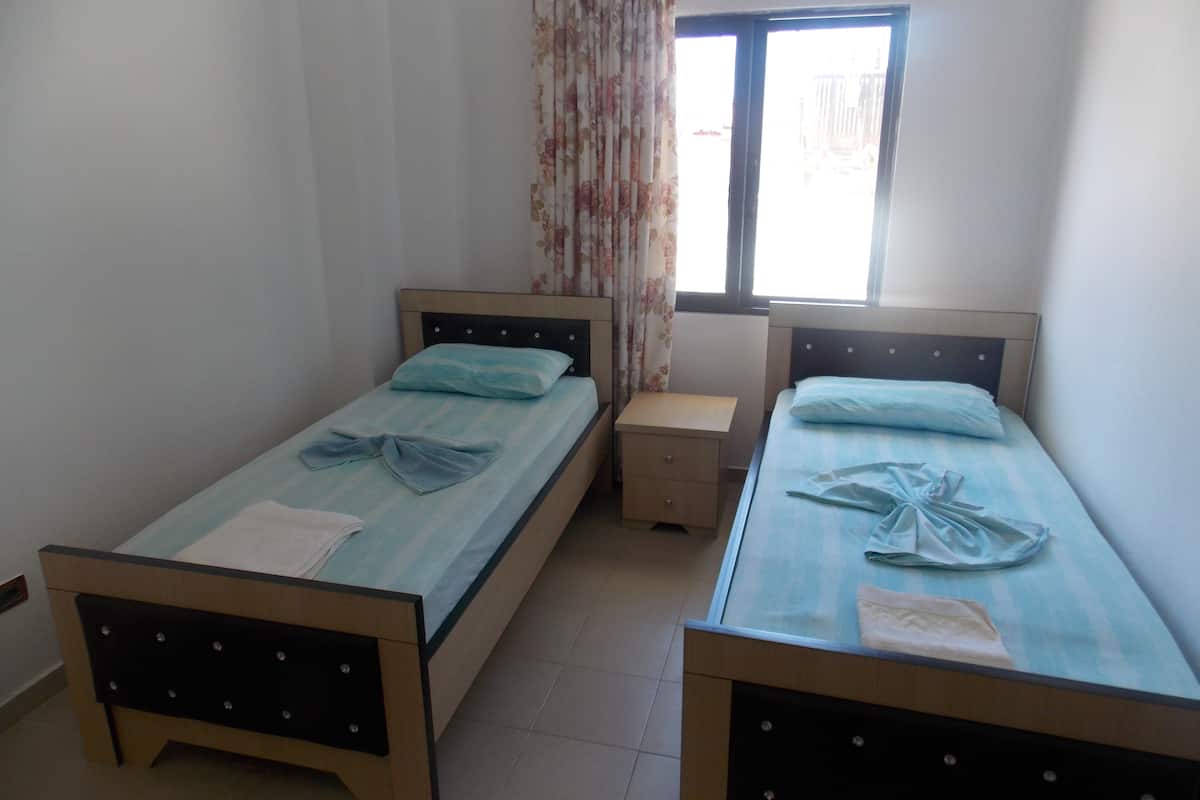 A room with two single beds, a small bedside table, a window with curtains, and a radiator.