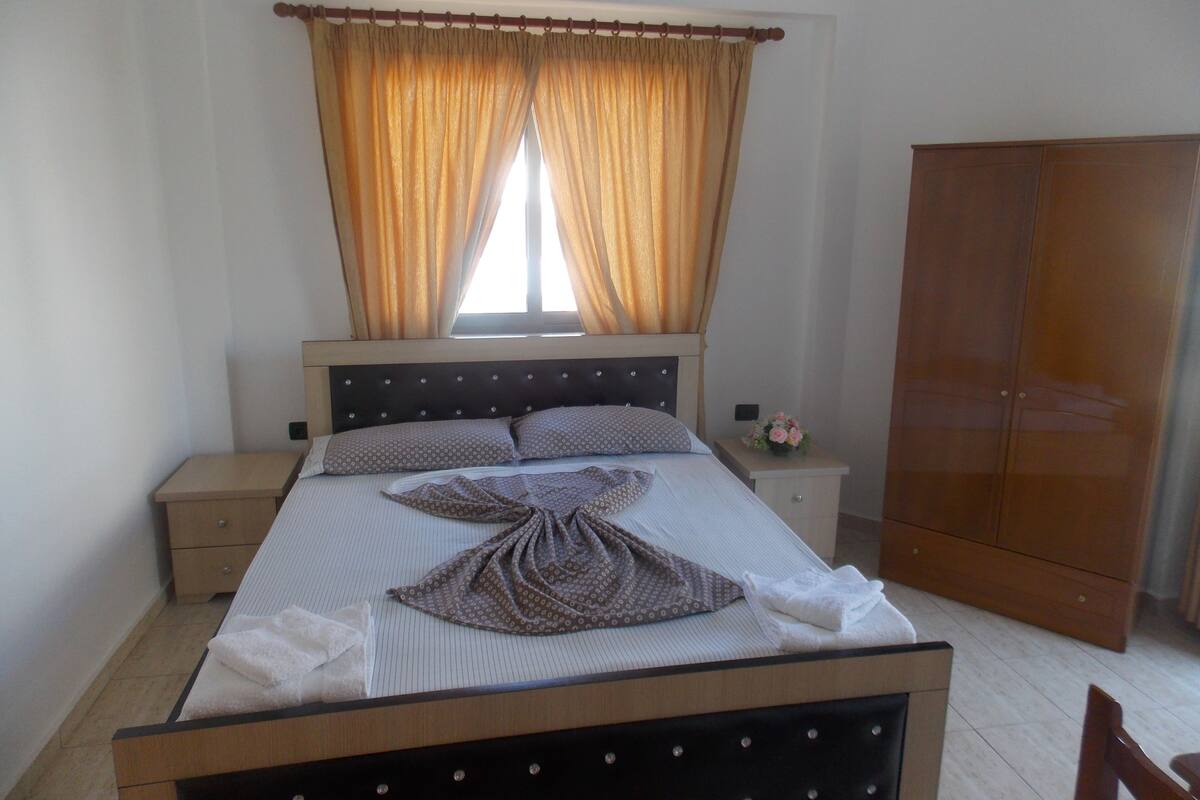 A neatly made bed with a striped bedspread, two pillows, and a decorative towel arrangement. There is a wooden nightstand with a small vase of flowers beside the bed. A wooden wardrobe is visible in the background.