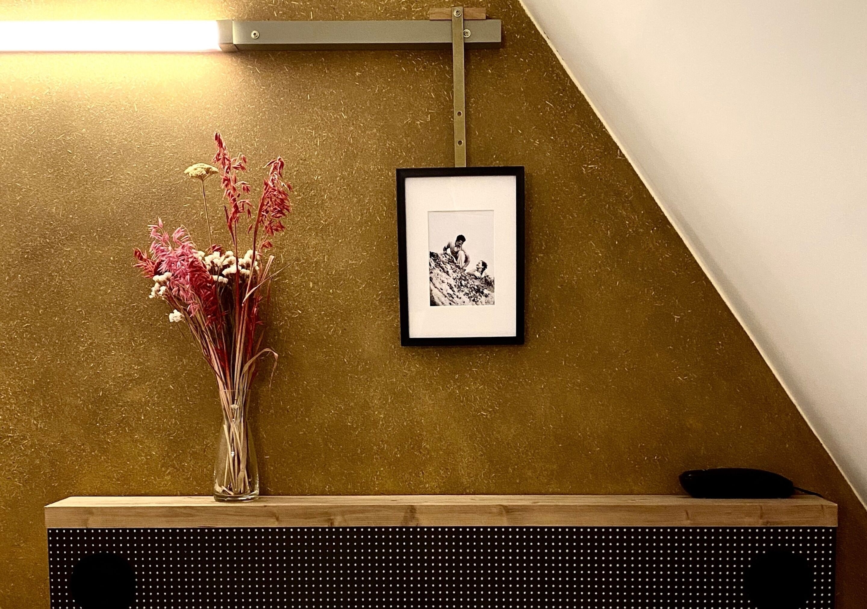 Una fotografía enmarco en la pared, un jarrón con flores en un estante y un escritorio de madera.