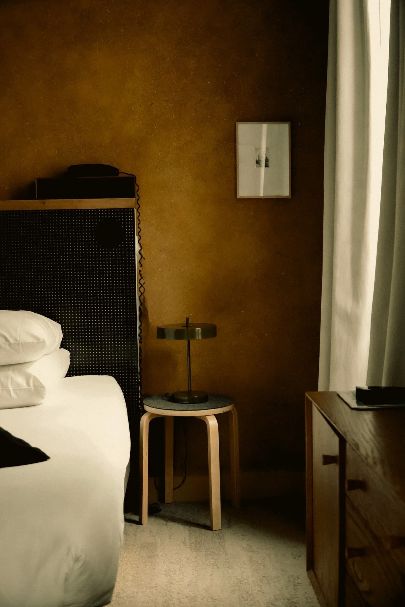 A hotel room with a bed, a small table with a lamp, a picture on the wall, and a wooden cabinet.
