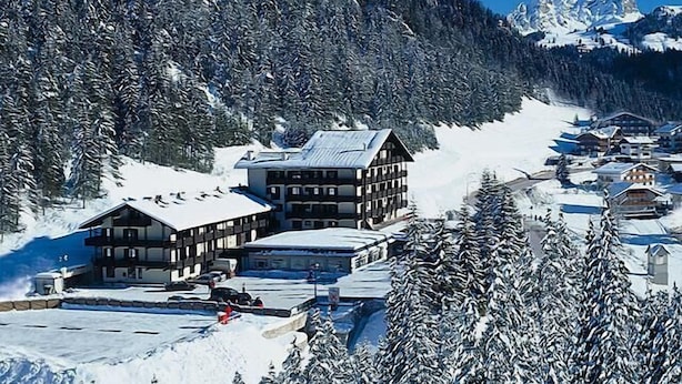A snow-covered mountain resort with multiple buildings, including a prominent lodge with a sloped roof, surrounded by dense forest and a clear blue sky. - Hotel Il Caminetto (Canazei)