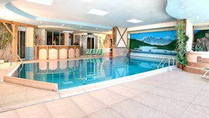 An indoor swimming pool with clear blue water, surrounded by a tiled floor and a stone wall. - Hotel Il Caminetto (Canazei)