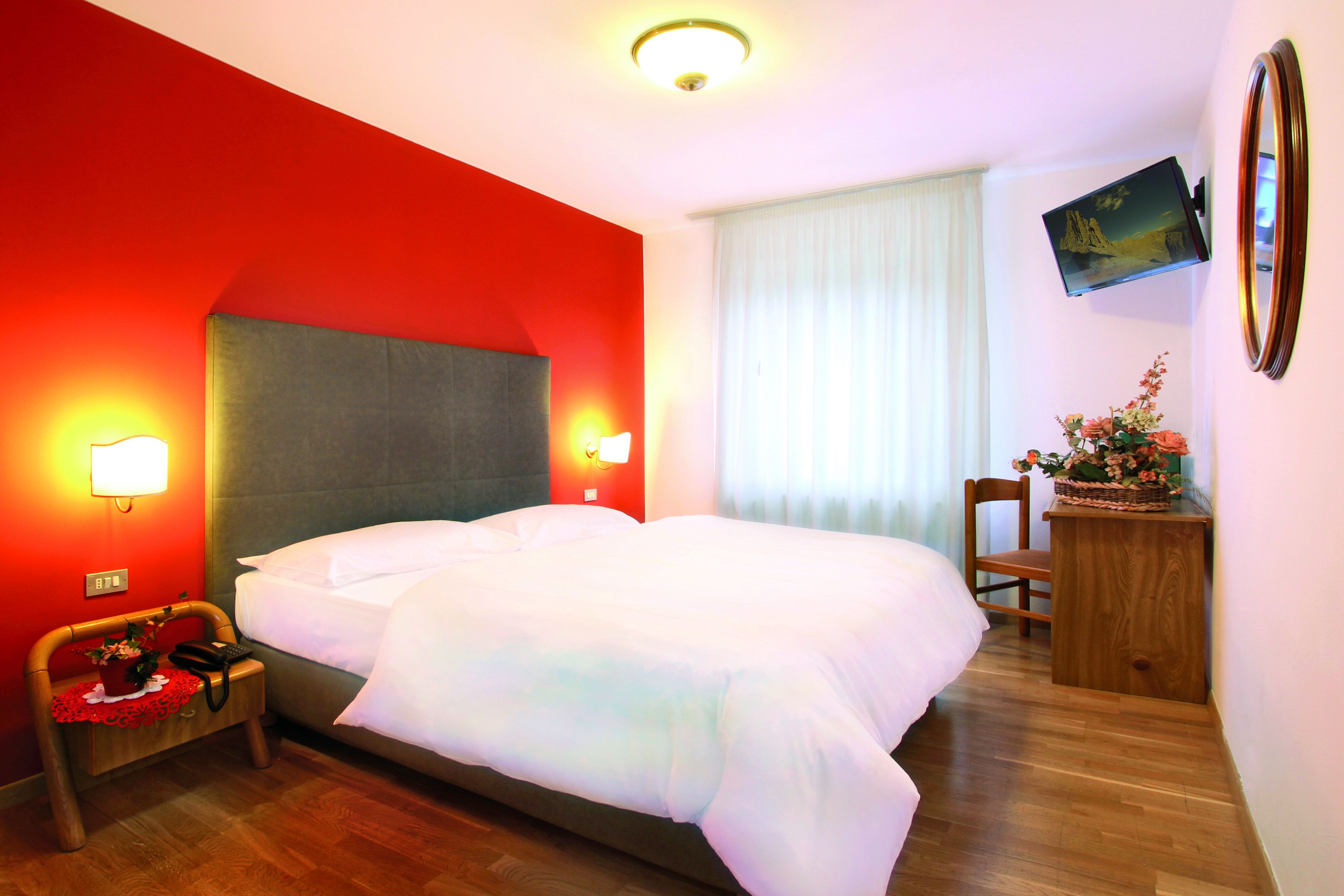 Habitació d'hotel amb un llit gran, dues làmpades de nit, una nitzeta de fusta amb un cist de flors, una cadira de fusta i un televisor de pantalla plana penjat a la paret.
