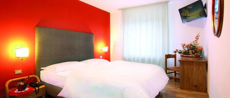 Habitació d'hotel amb un llit gran, dues làmpades de nit, una nitzeta de fusta amb un cist de flors, una cadira de fusta i un televisor de pantalla plana penjat a la paret.