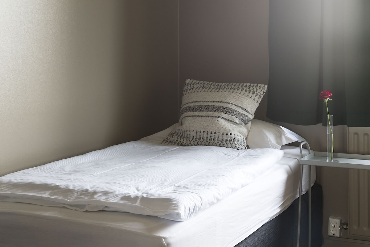 A neatly made bed with pillows, a bedside table with a vase of flowers, and a radiator under the window.