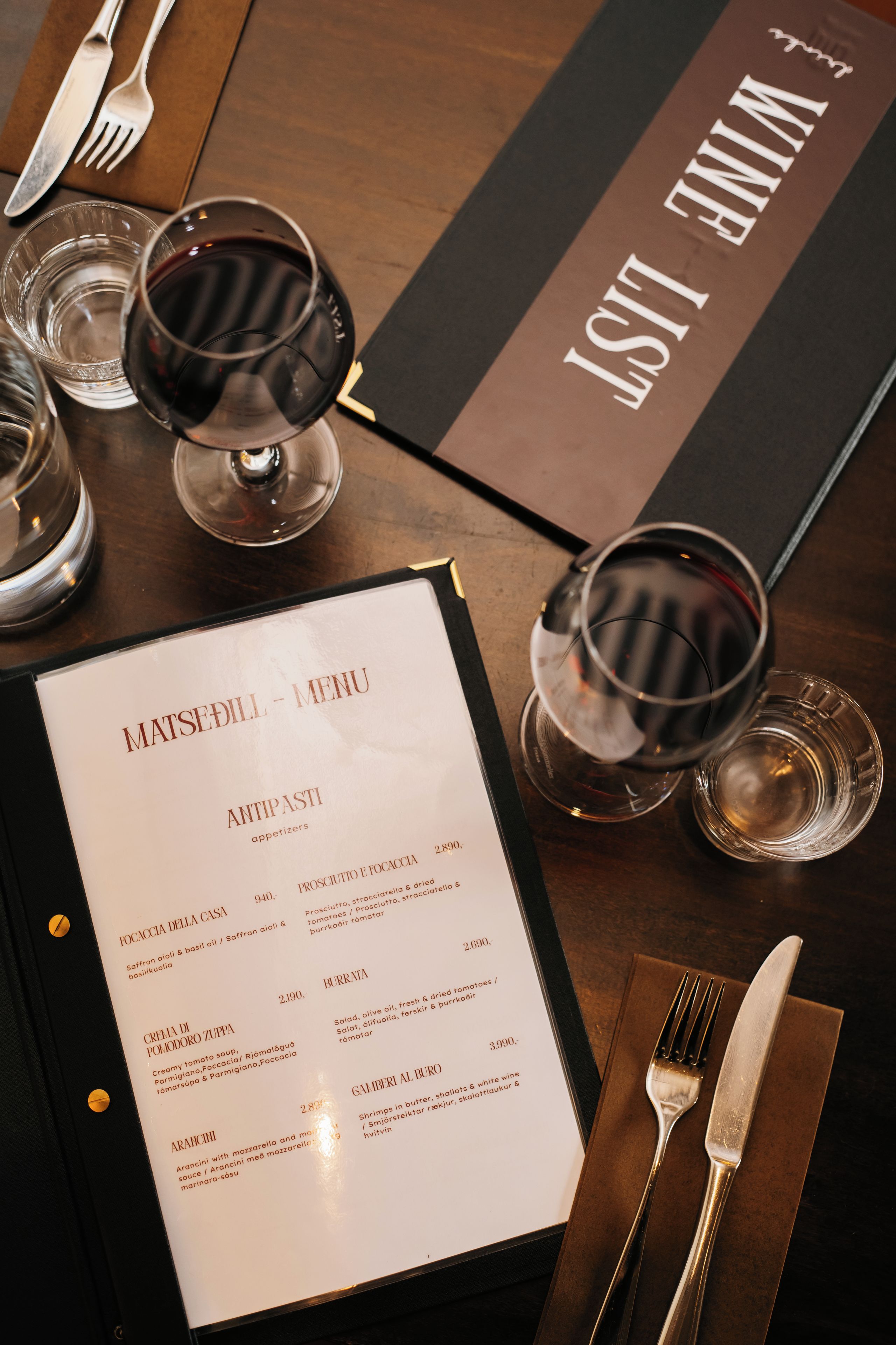 A menu with various dishes and their prices, a wine glass, and cutlery on a table.