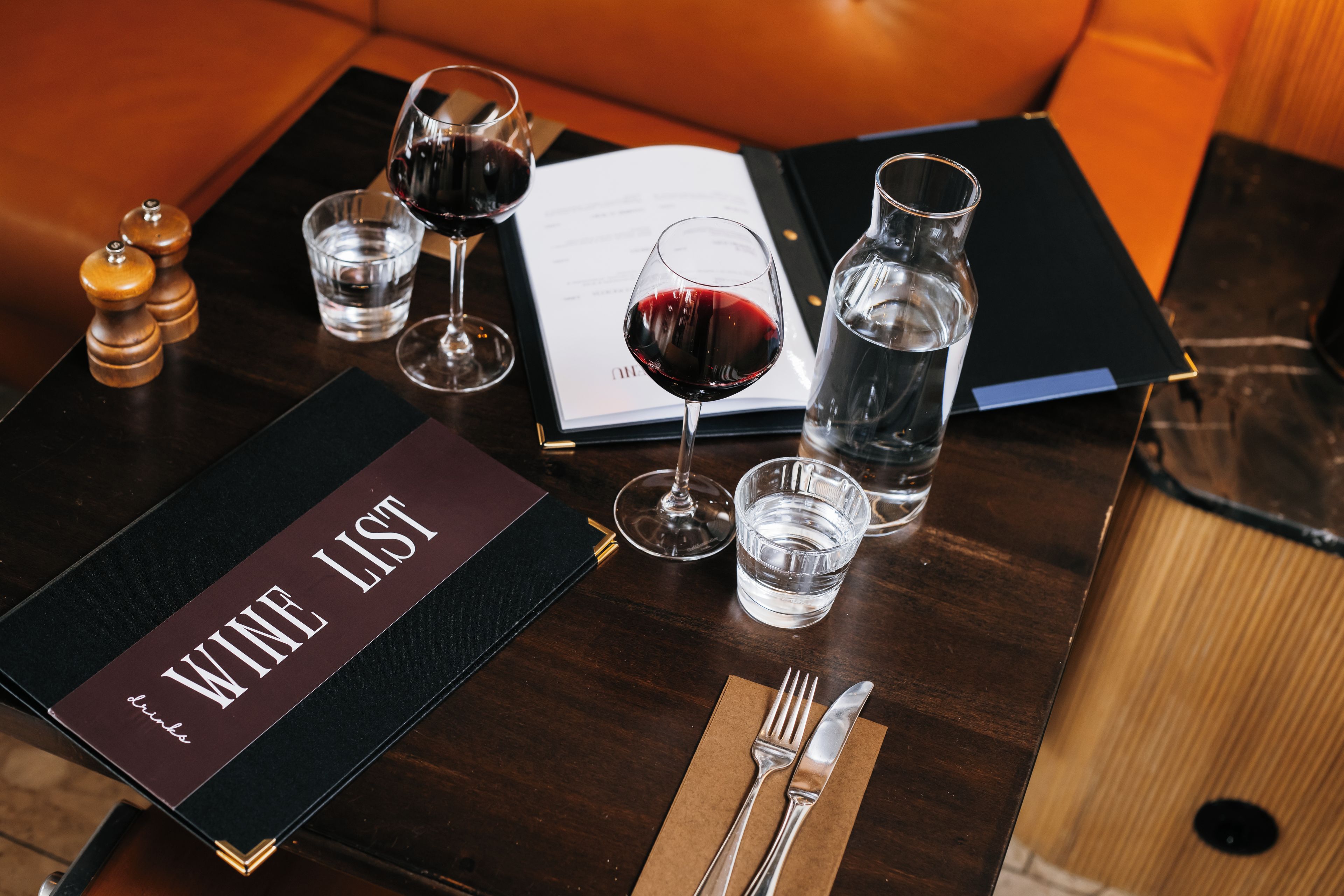 A table with a wine list, glasses, water, a menu, and cutlery.
