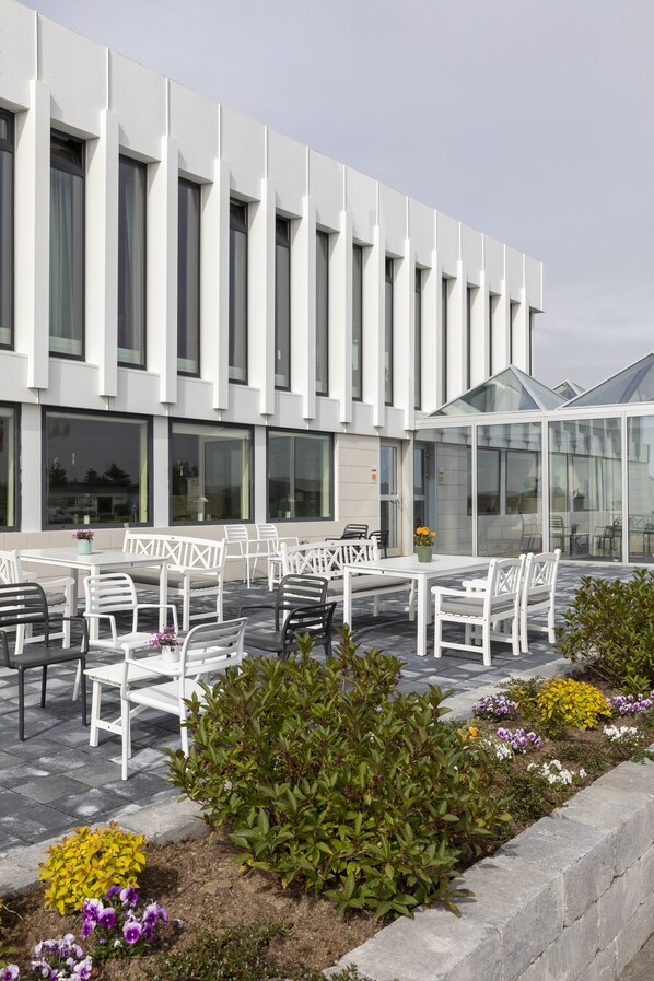 A modern building with a white facade, large windows, and outdoor seating with white chairs and tables. - Hotel Höfn (Höfn)