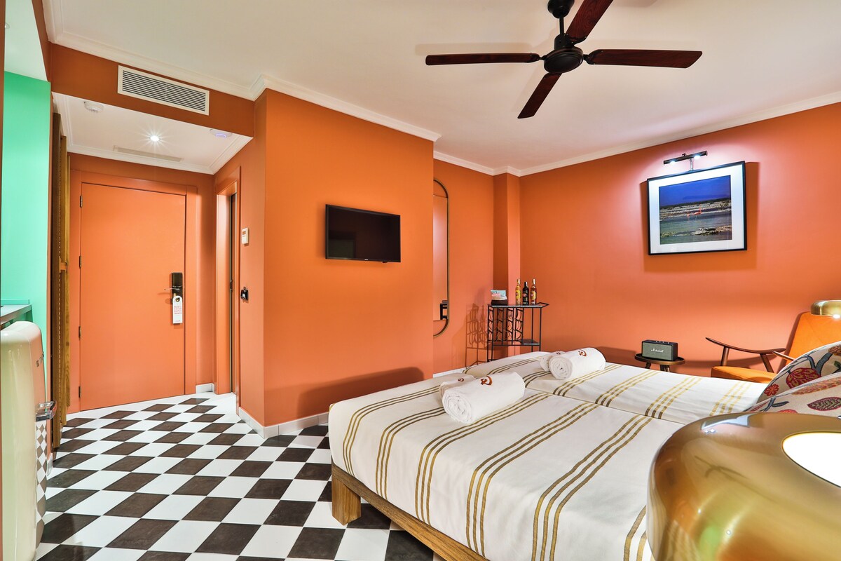 Een hotelkamer met een geruit vloerkleed, een bed met wit-goud gestreepte beddengoed, een plafondventilator, een wandgemonteerde televisie en een ingelijste foto aan de muur.