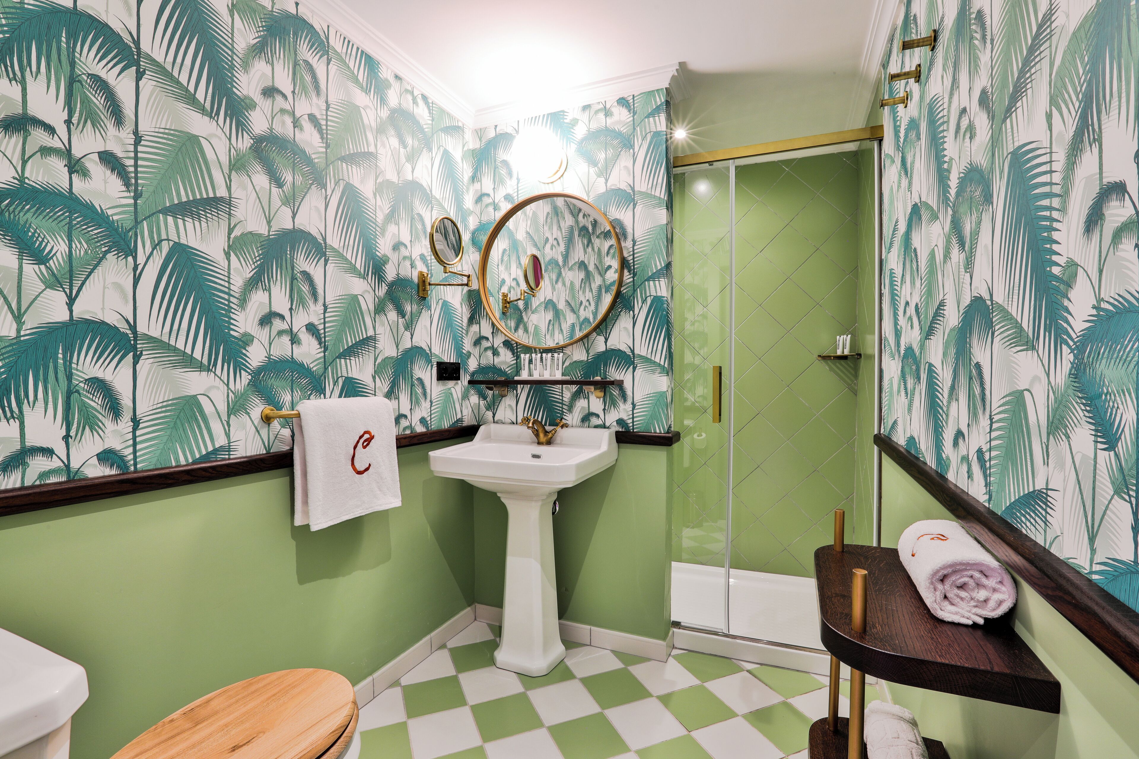 A bathroom with a green and white checkered floor, a white pedestal sink, a wooden toilet, a glass shower enclosure, and a mirror with a gold frame.
