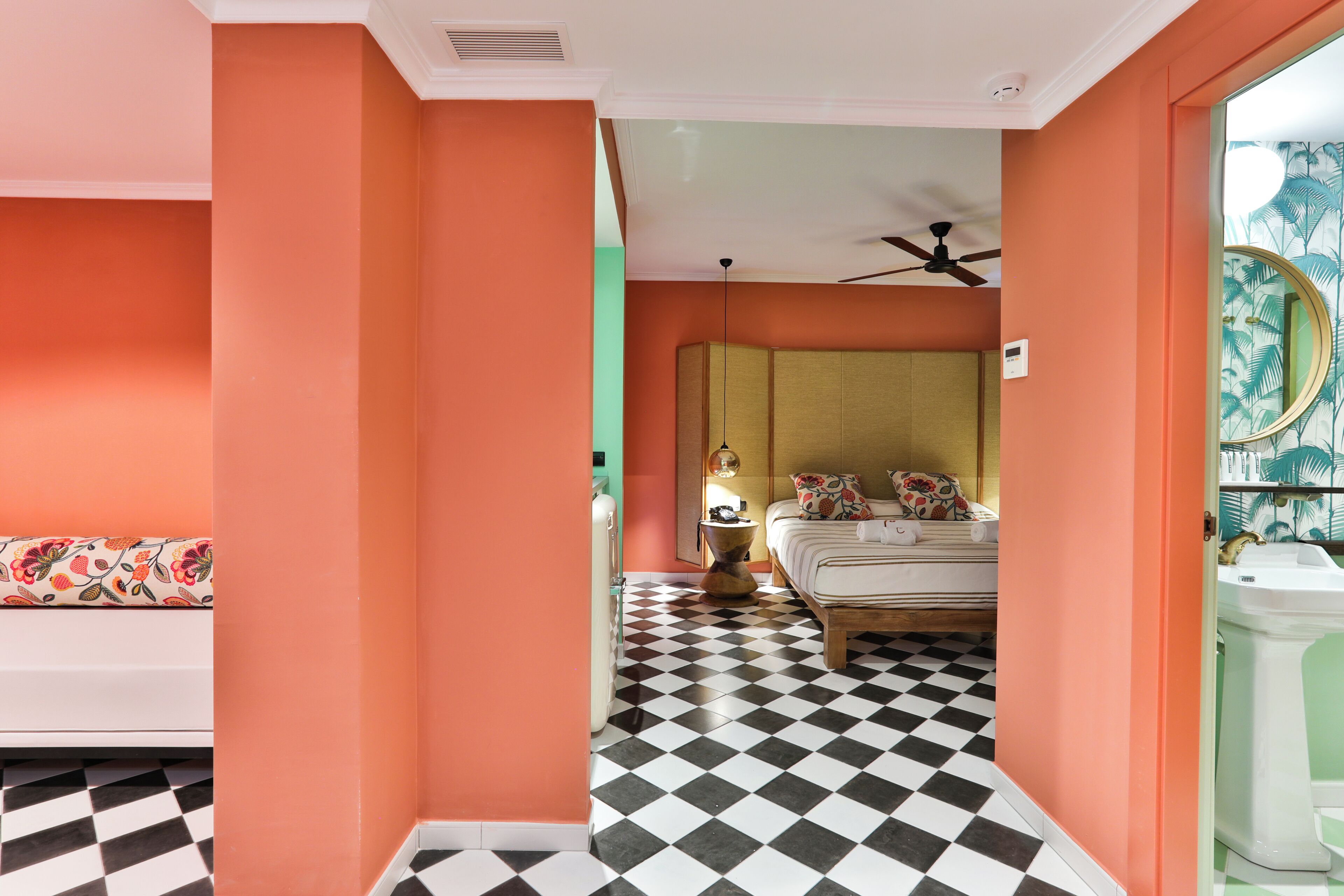 A hotel room with a checkered floor, a bed with floral pillows, a white sofa, and a bathroom with a green wall and a round mirror.