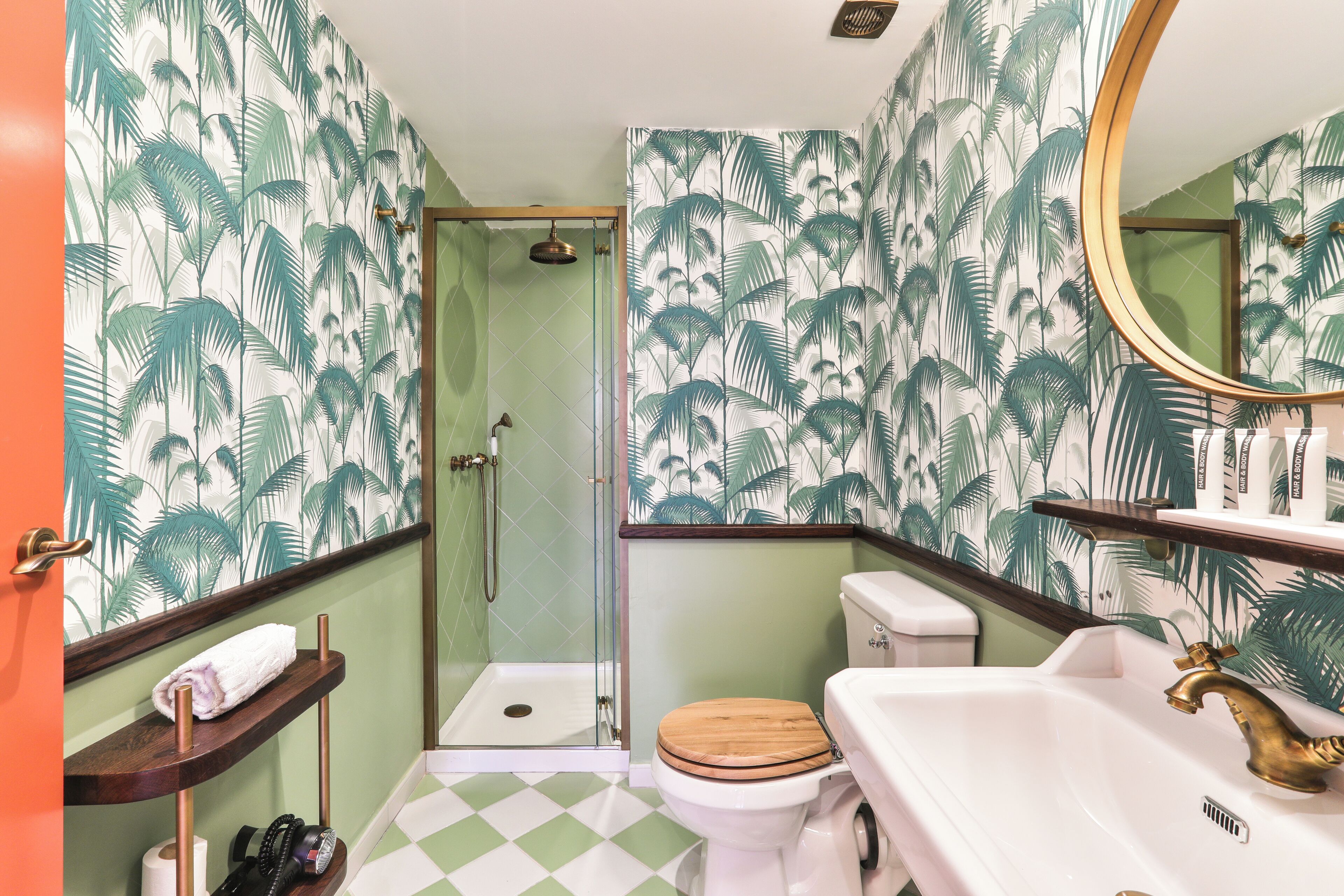 Une salle de bain équipée d’une douche, de toilettes et d’un lavabo, avec un papier peint à motifs tropicaux et un carrelage à carreaux.