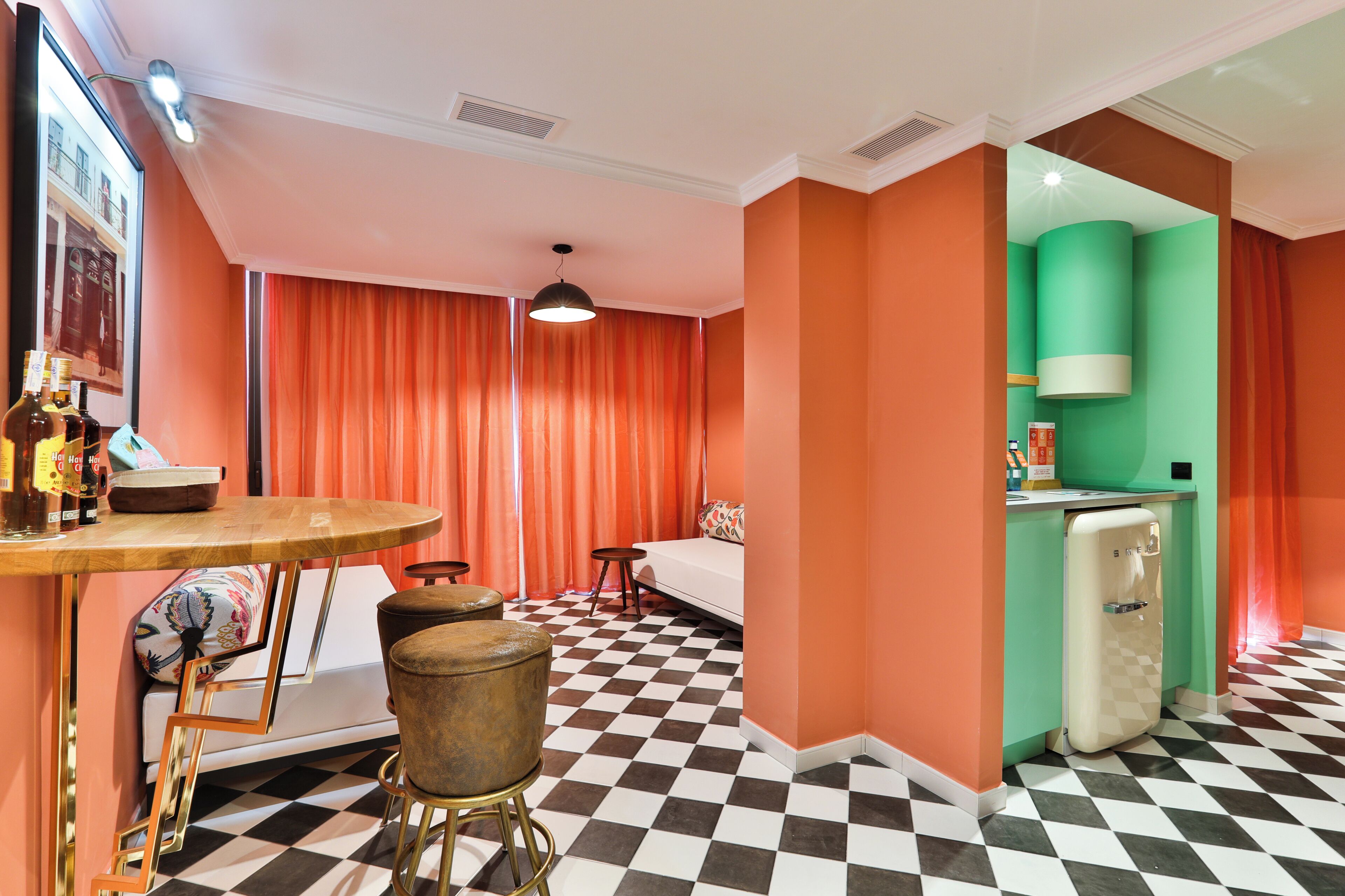 A room with a checkered floor, a sofa, a table with stools, a refrigerator, and a wall-mounted light fixture.
