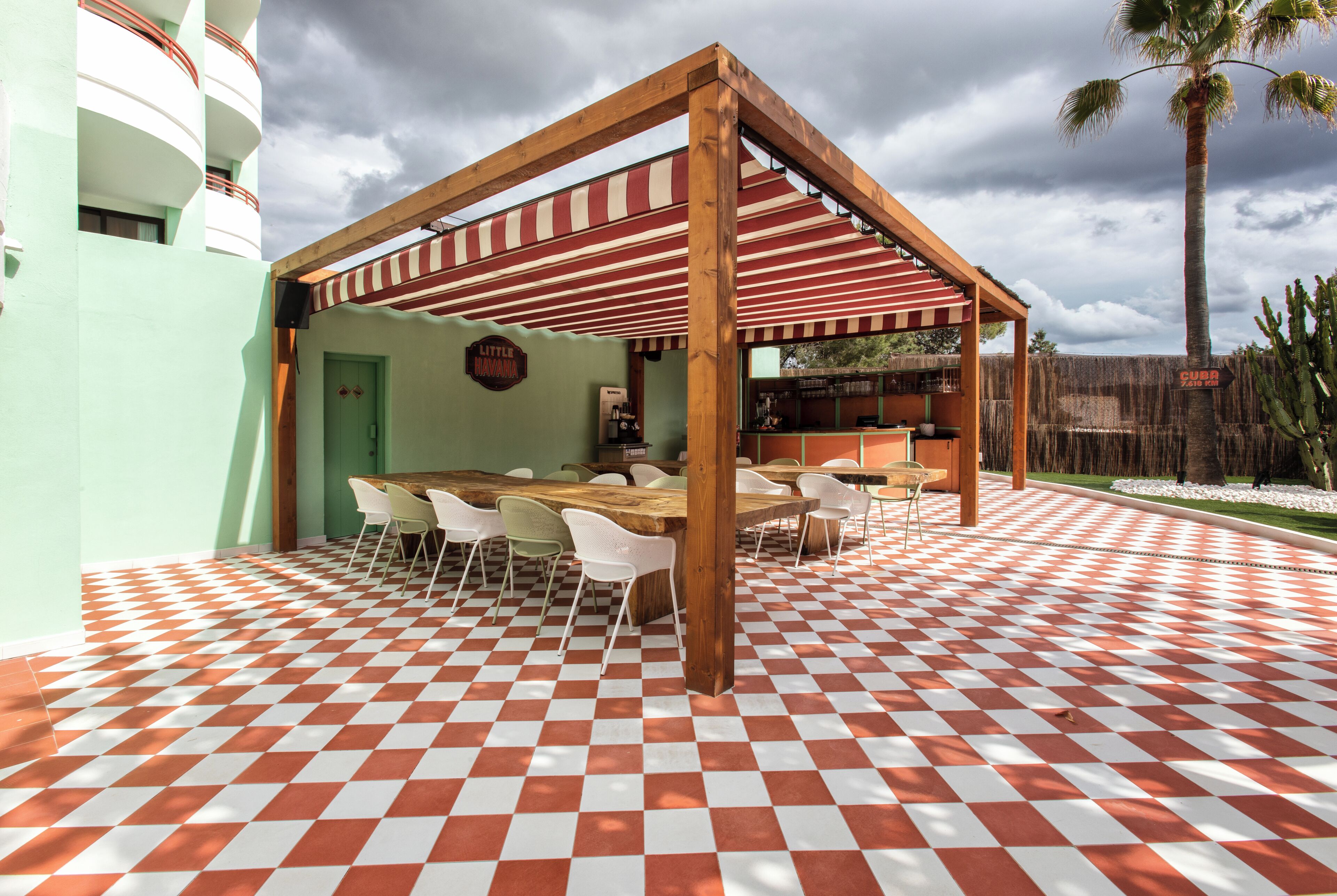 Een overdekt terras met een geruit vloertje, houten tafels en stoelen.