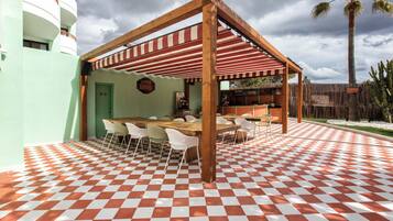 Een overdekt terras met een geruit vloertje, houten tafels en stoelen.