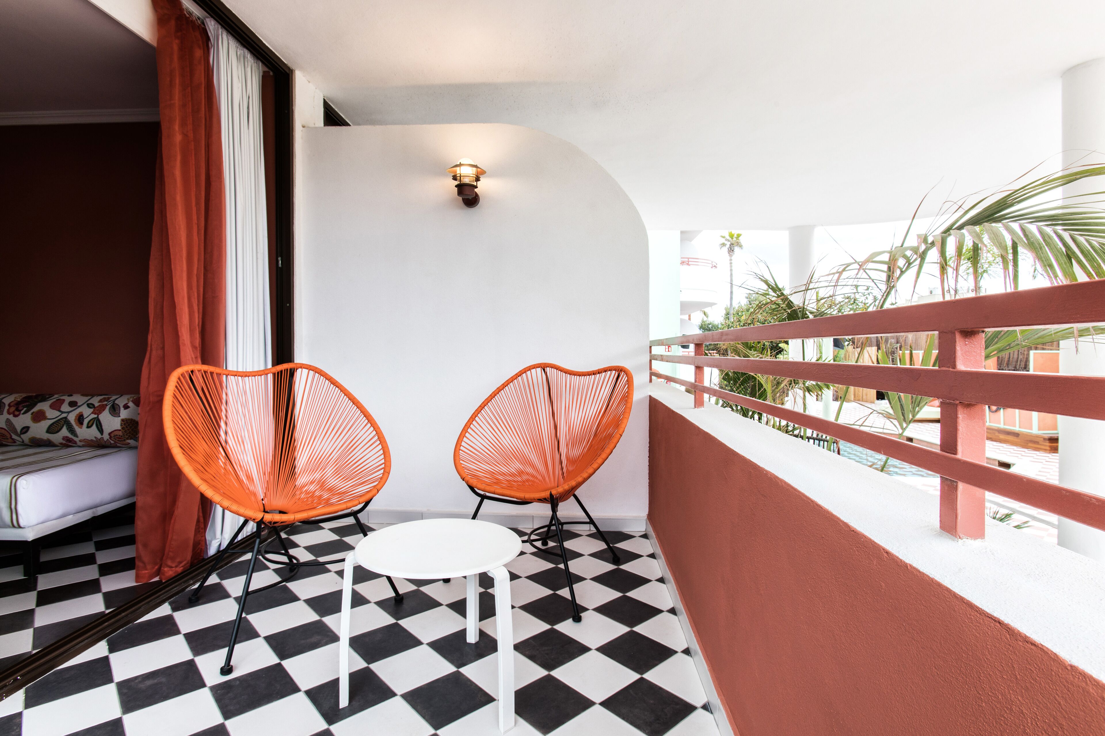 A balcony with a checkered floor, two orange chairs, a white table, and a sofa with a floral pattern.