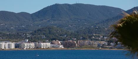 A coastal cityscape with buildings along the shore, a body of water, and a mountain range in the background.