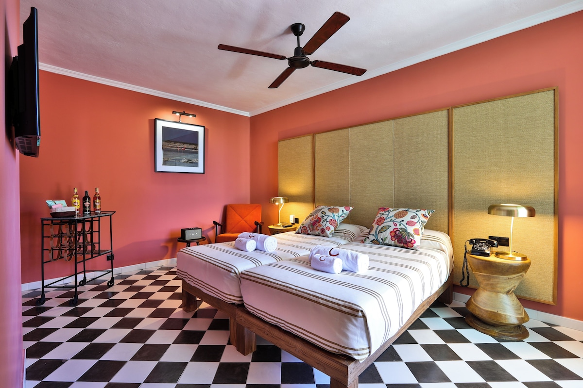 Ein Hotelzimmer mit Schachbrettboden, einem Bett mit gestreifter Bettwäsche, einem Deckenventilator, einem Nachttisch mit Lampe und einem an der Wand montierten Fernseher.