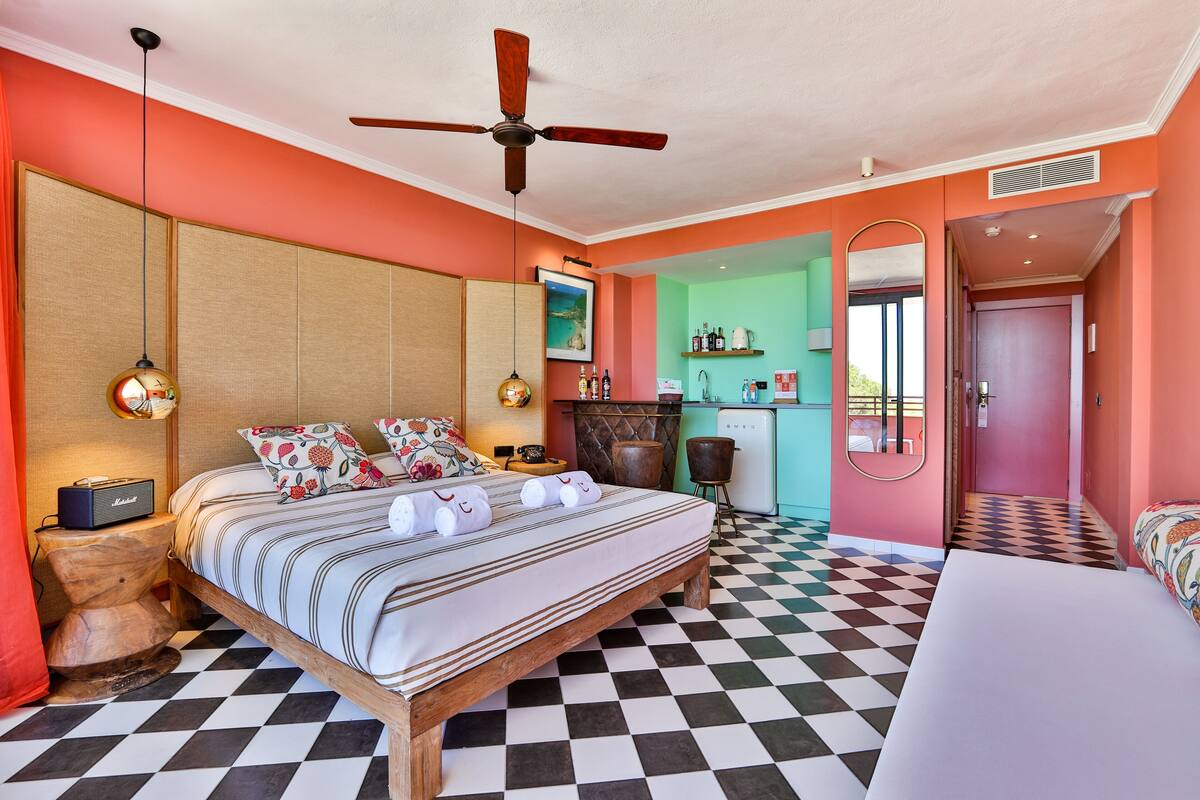 Une chambre d’hôtel avec un sol à carreaux, un lit avec une literie à rayures, un ventilateur de plafond, un minibar et une vue sur l’extérieur.