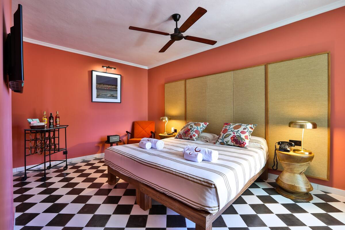 Une chambre d’hôtel avec un sol à carreaux, un lit avec une literie à rayures, un ventilateur de plafond, une table de chevet avec une lampe et un téléviseur fixé au mur.