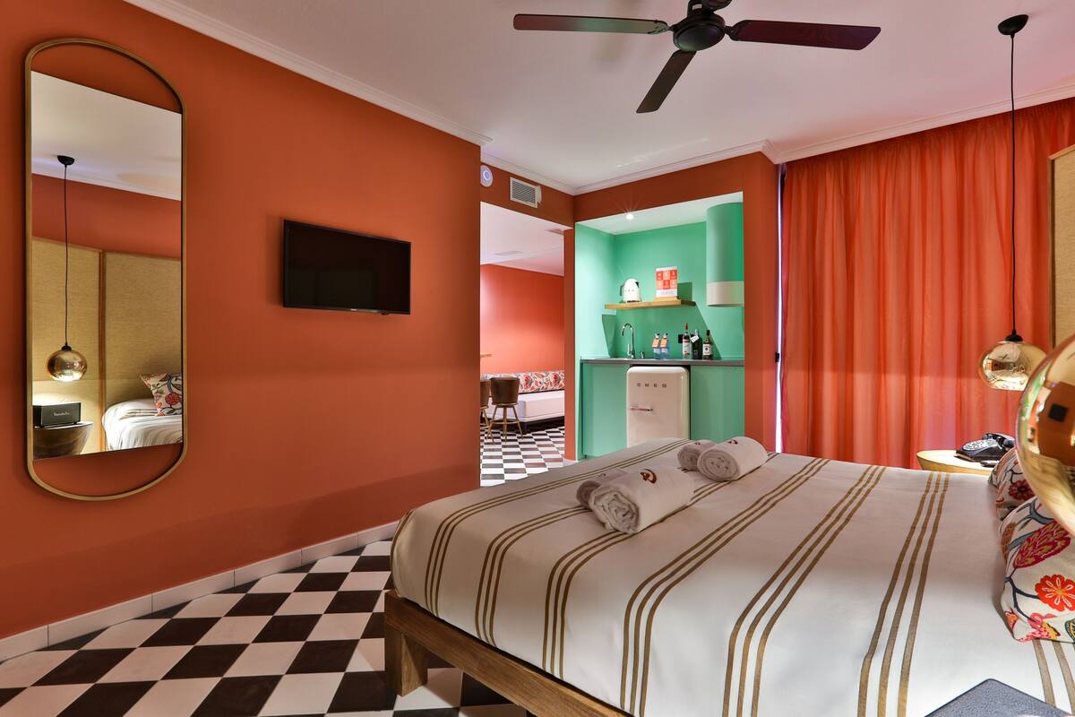 Une chambre d’hôtel avec un sol à carreaux, un lit avec une literie à rayures, un ventilateur de plafond, une télévision et un mini-bar.