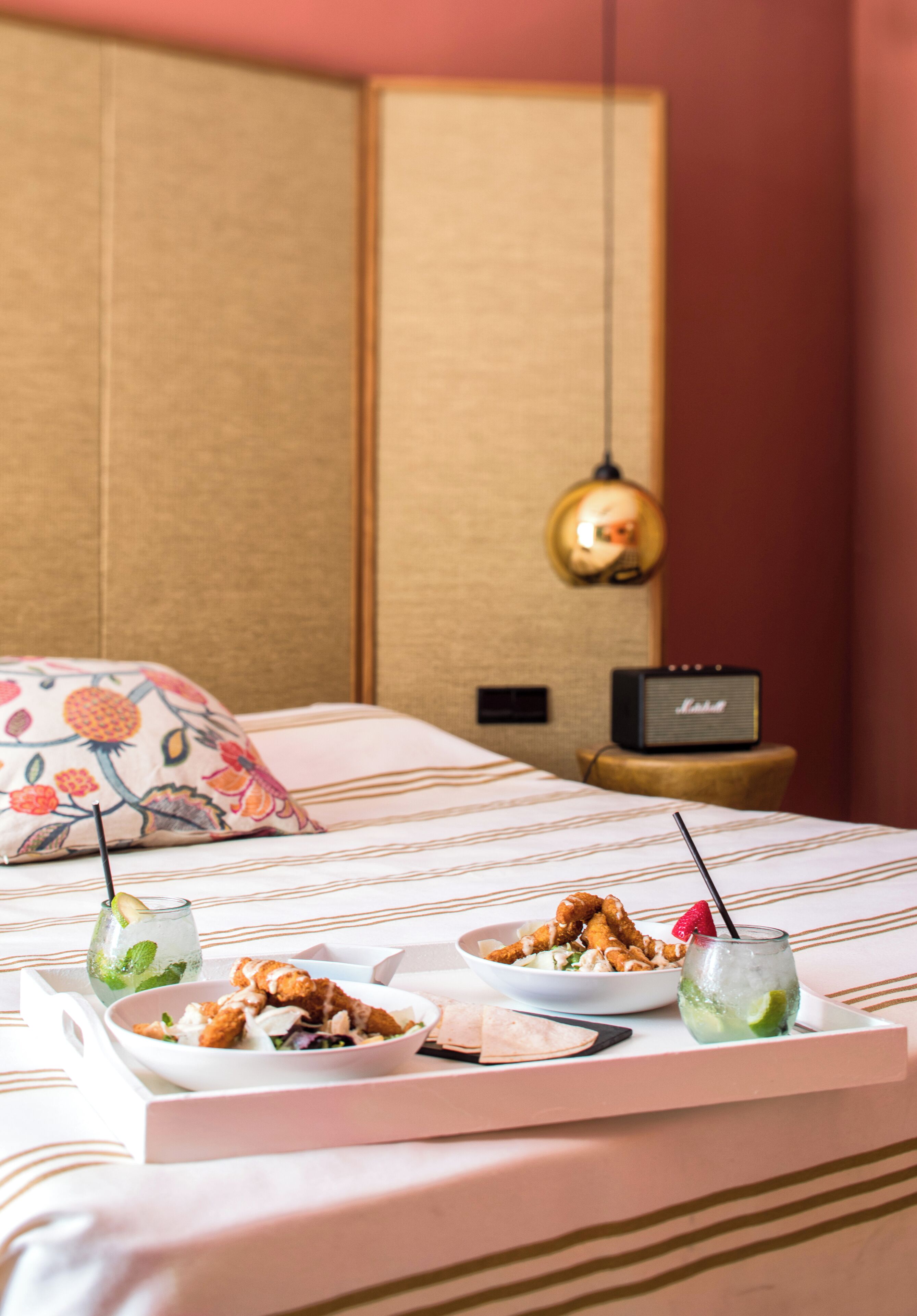 A neatly made bed with a floral pillow, a small speaker, and a tray with food and drinks.