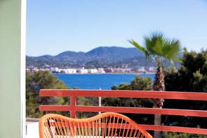 Un balcon offrant une vue sur une ville côtière, un palmier et des montagnes à l’horizon.