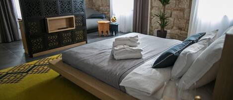 A modern hotel room with a large bed, a flat-screen TV mounted on a decorative wall, a small seating area with a sofa and side table, and a stone wall in the background.