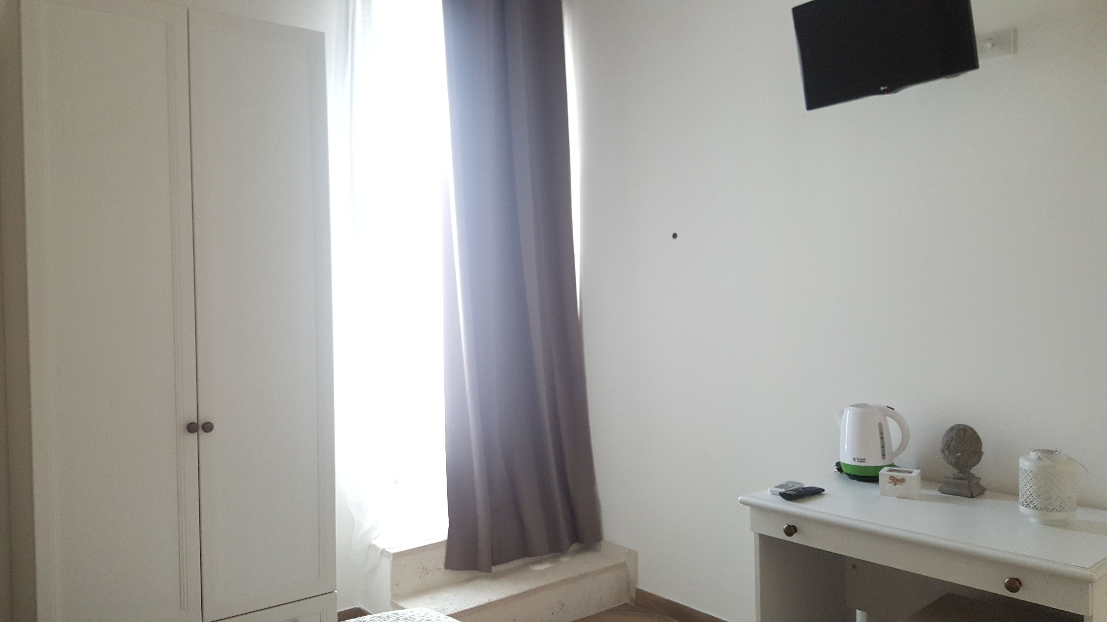 A room with a white wardrobe, a window with a grey curtain, a white desk with a kettle, a remote control, and a small statue.