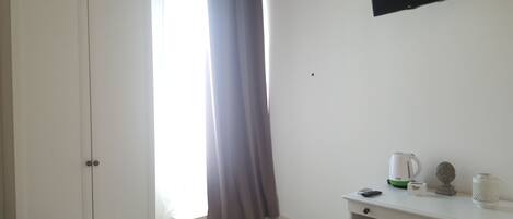 A room with a white wardrobe, a window with a grey curtain, a white desk with a kettle, a remote control, and a small statue.