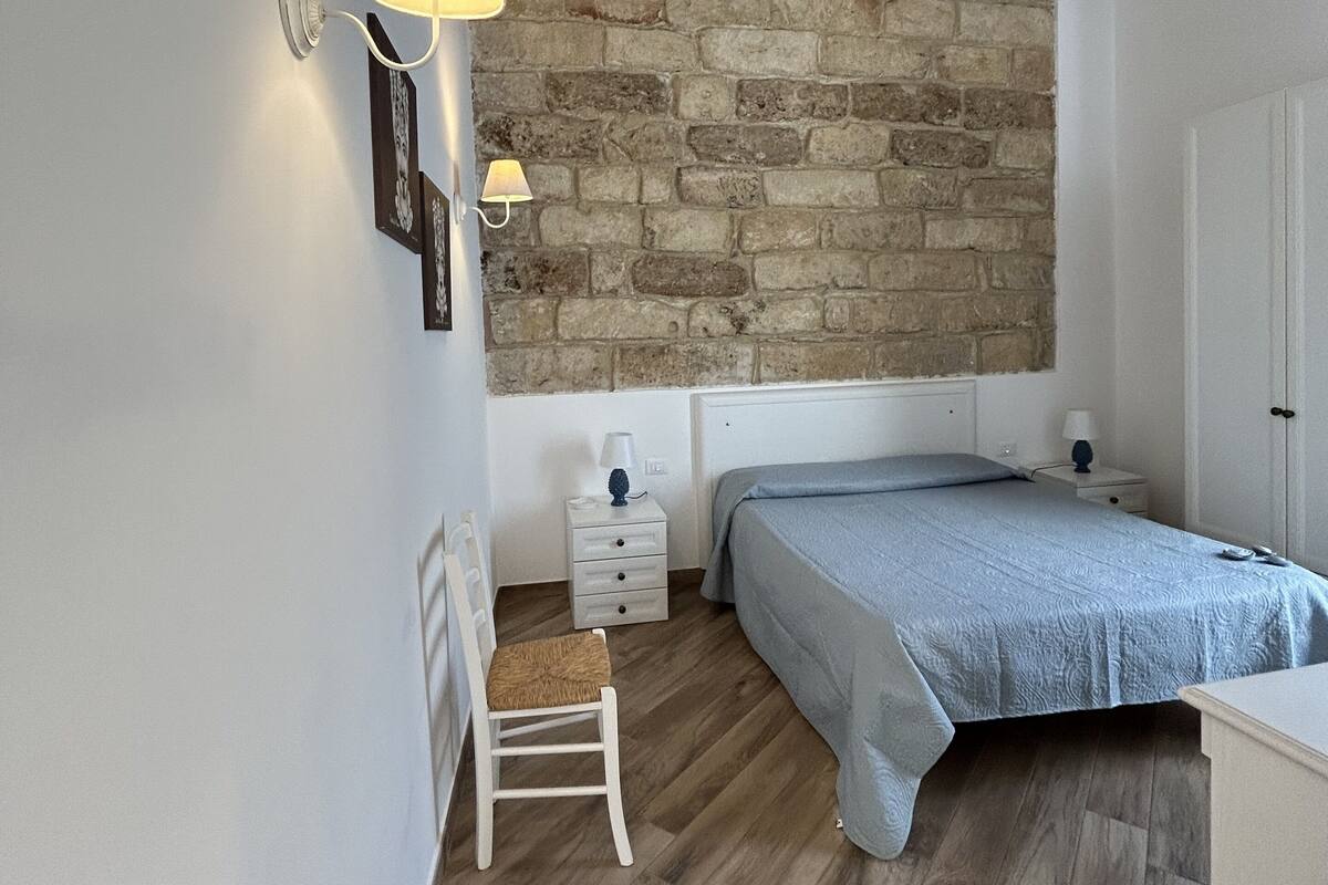 Quarto com parede de pedra, soalho de madeira, cama, mesa de cabeceira, cadeira e abajur.