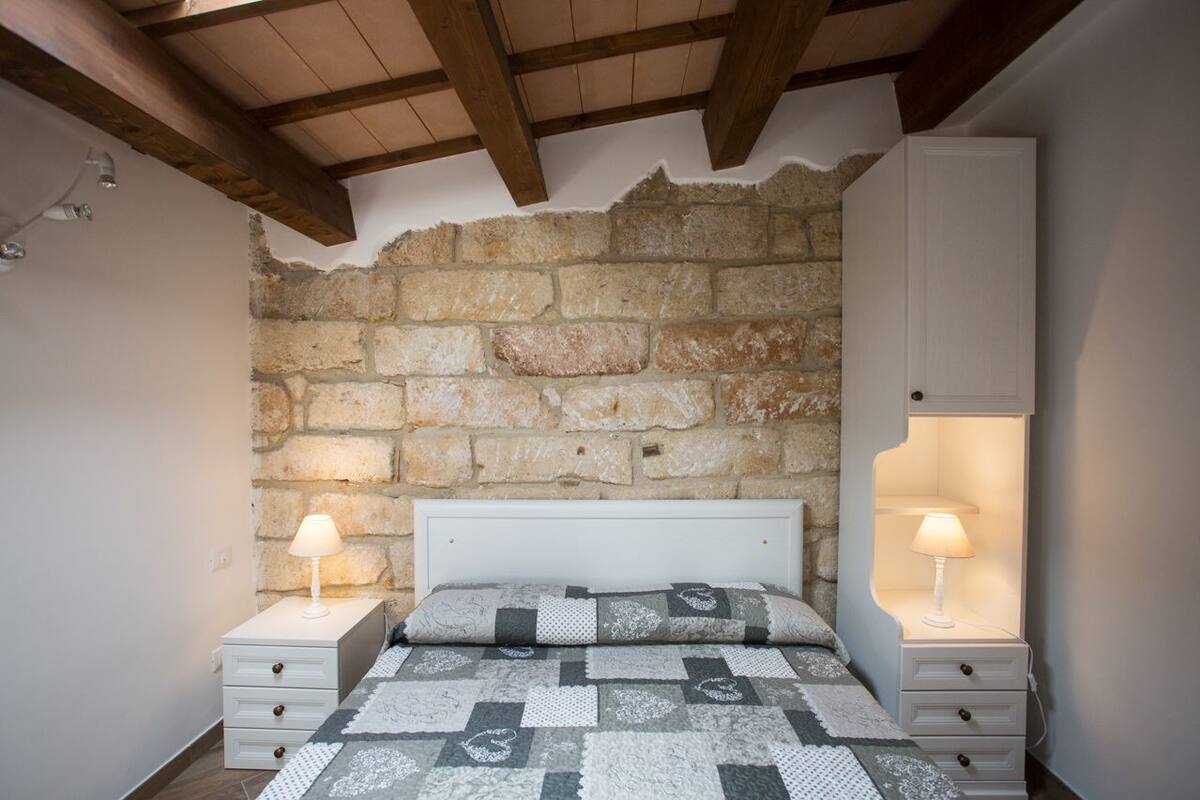 Quarto com parede de pedra, cama com colcha estampada, duas mesas de cabeceira com abajures e cabeceira branca.