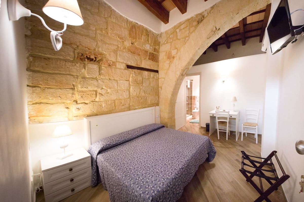 Quarto com parede de pedra, cama com colcha estampada, escrivaninha com cadeira e mesa pequena com abajur.