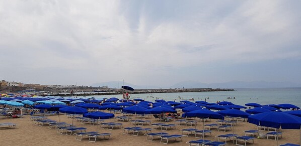 Una playa con numerosas sombrillas azules y sillas de playa, un espigón rocoso, y vistas al mar y montañas distantes.